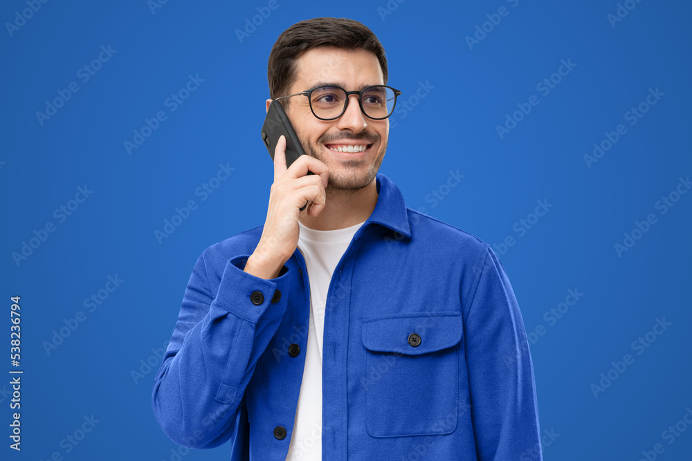 Portrait of handsome man in blue shirt and glasses, answering phone call, looking aside with smile Portrait of handsome man in blue shirt and glasses, answering phone call, looking aside with smile