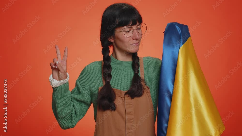Young nerd woman with pigtails in glasses holding Ukraine national flag, showing victory sign against war, hoping for success and win. Girl doing peace gesture, smiling with kind optimistic expression