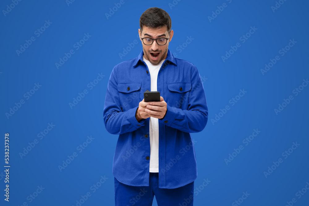 Surprised man in blue shirt looking at phone with amazed face expression, shocked by new online app Surprised man in blue shirt looking at phone with amazed face expression, shocked by new online app