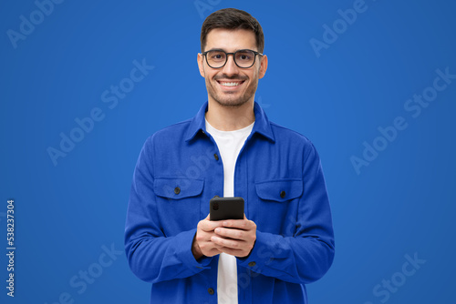 Young modern man in casual blue shirt and glasses, holding smart phone in hands, looking at camera
