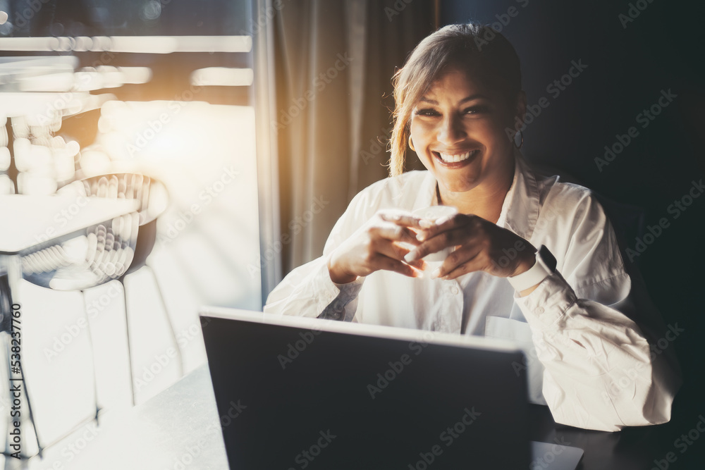 Portrait of a happy smiling Hispanic female entrepreneur in a white shirt, sitting with a cup of expresso coffee in her hands in front of her laptop next to the window of a cafe or a restaurant - obrazy, fototapety, plakaty 