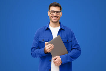 Young smiling modern male teacher holding laptop on blue background