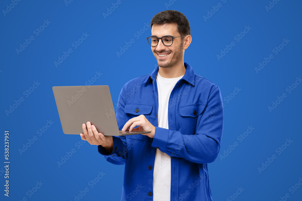 Young stylish man wearing casual blue shirt, standing with opened laptop, surfing online or typing