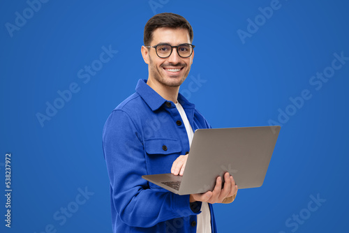 Portrait of modern business man standing in casual blue shirt, holding laptop and looking at camera