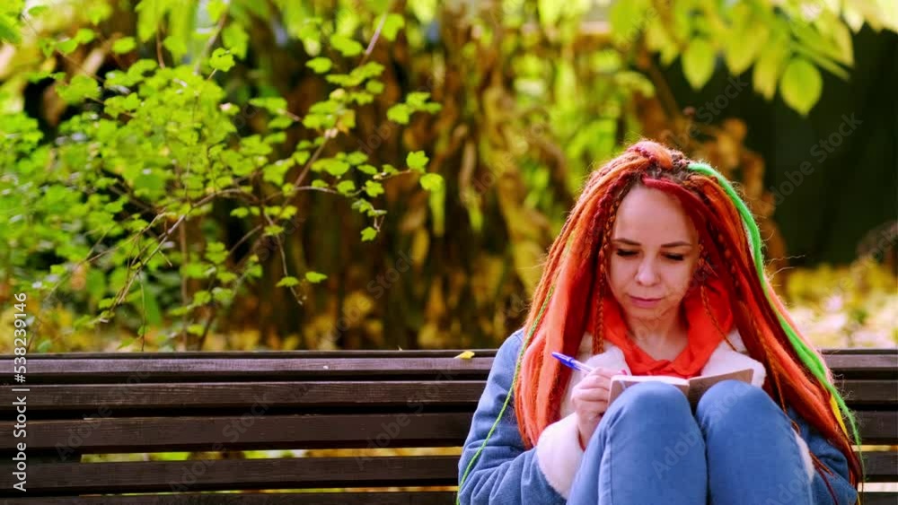 Young woman student with long hair in casual clothes sitting on wooden bench and writing notes in notebook Young woman student with long hair in casual clothes sitting on wooden bench and writing notes in notebook