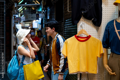 Young Asian couple enjoy and fun outdoor lifestyle shopping together at street market on summer holiday vacation. Happy man and woman choosing and buying fashion clothes together at clothing shop.