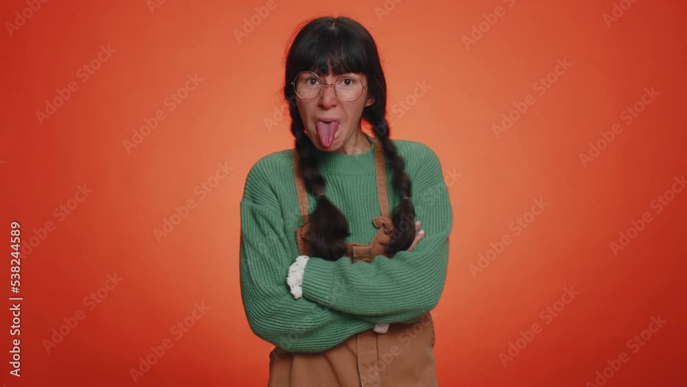 Cheerful funny woman in green sweater showing tongue making faces at camera, fooling around, joking, aping with silly face teasing. Young nerd adult girl isolated alone on orange studio background