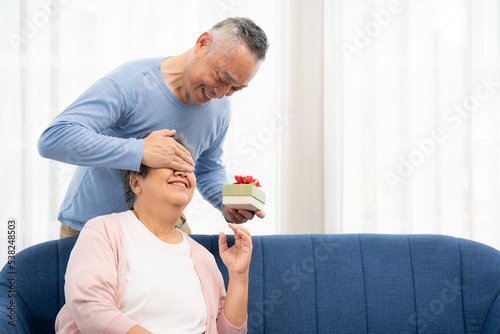 Happy Senior Asian man covering eyes of senior Asian woman for surprise with gift box in home. Elderly Asian couple on valentine's day concept.