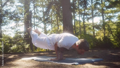 Close up shot caucasian adult man dressed in white holds an advanced yoga pose, peacock mayurasana. In pine tree forest, camera movement, handheld. photo