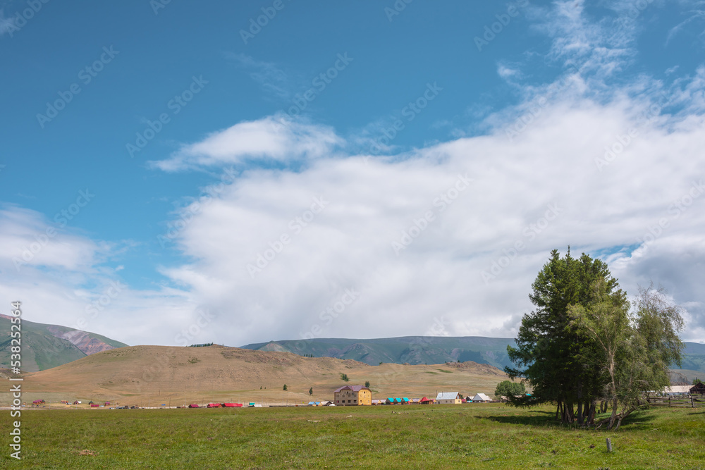 Dramatic alpine view from trees to small houses in sunlit green meadow against high mountain range under clouds in blue sky. Scenic mountain landscape at changeable weather. Village in high mountains. Dramatic alpine view from trees to small houses in sunlit green meadow against high mountain range under clouds in blue sky. Scenic mountain landscape at changeable weather. Village in high mountains.