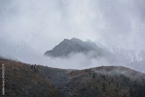 Dark atmospheric landscape with high mountain silhouettes in dense fog in rainy weather. Snowy rocky mountain top above hills in thick fog in dramatic overcast. Black rocks in low clouds during rain.