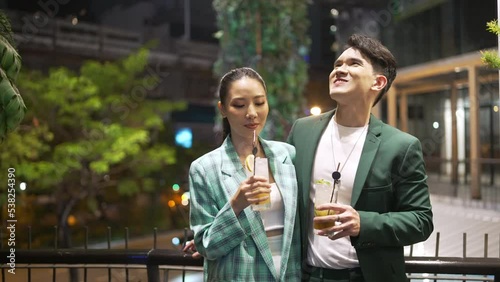 4K Happy Asian couple celebrating holiday event at luxury restaurant in the city at night. Attractive man and woman enjoy city lifestyle drinking cocktail together at bar terrace on travel vacation.
