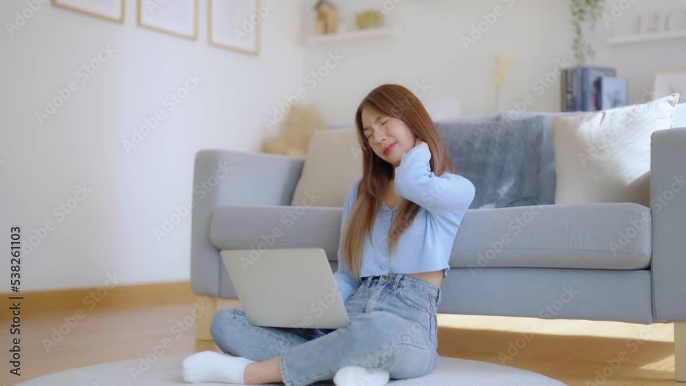 Office syndrome concept. Asian young woman working on laptop while seated on floor next to couch at home, feeling pain in neck and shoulder after working on computer laptop for a long time Office syndrome concept. Asian young woman working on laptop while seated on floor next to couch at home, feeling pain in neck and shoulder after working on computer laptop for a long time