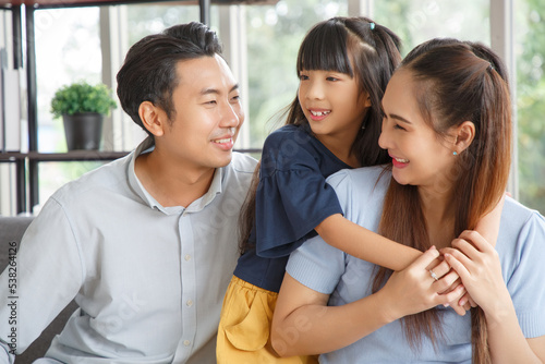 Portrait of a happy young family. Mom, dad and daughter look each other and smile. The faces of Asian parents and their child in living room.