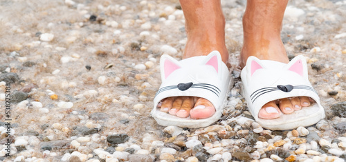 Legs of a girl by the sea. Feet in funny slippers on pebbles. Sea wave and slippers. Slippers in the form of kittens.