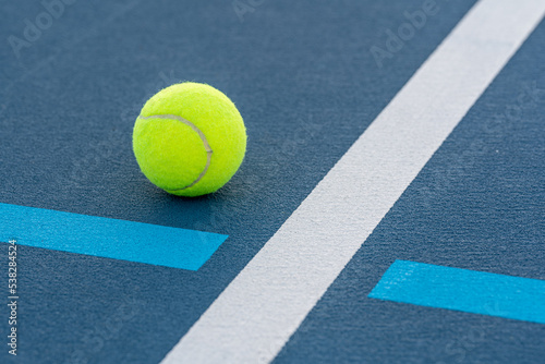 Photo taken in late evening under lights of a yellow tennis ball on blue tennis court with white line and green out of bounds. © Thomas