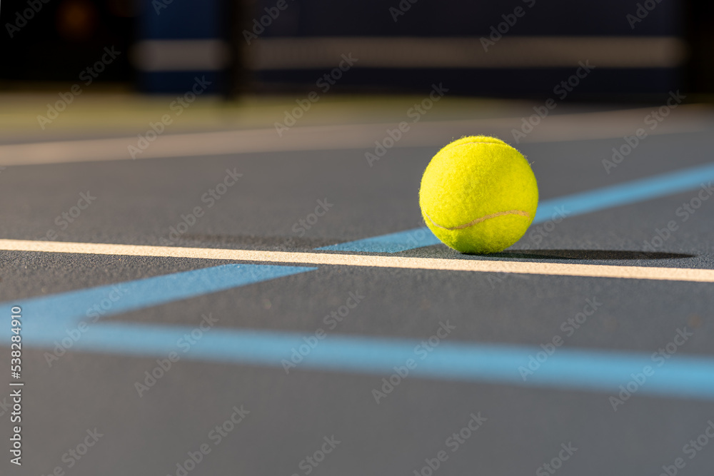 Photo taken in late evening under lights of a yellow tennis ball on blue tennis court with white line and green out of bounds.