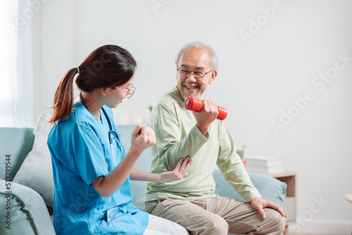 Asian Senior old man doing physiotherapist with support from caregiver. Mature male sitting on sofa in living room using dumbbell workout exercise. Healthcare medical concept.
