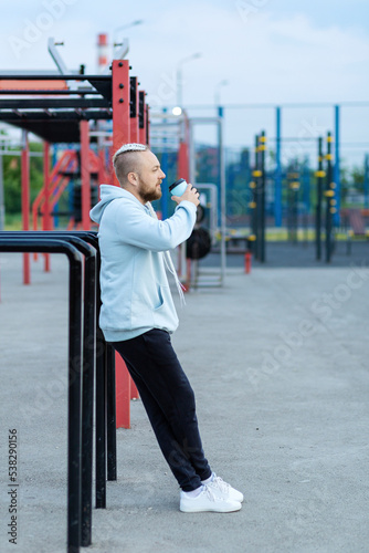 A man with a braid hairstyle in the early morning drinks coffee on the sports ground