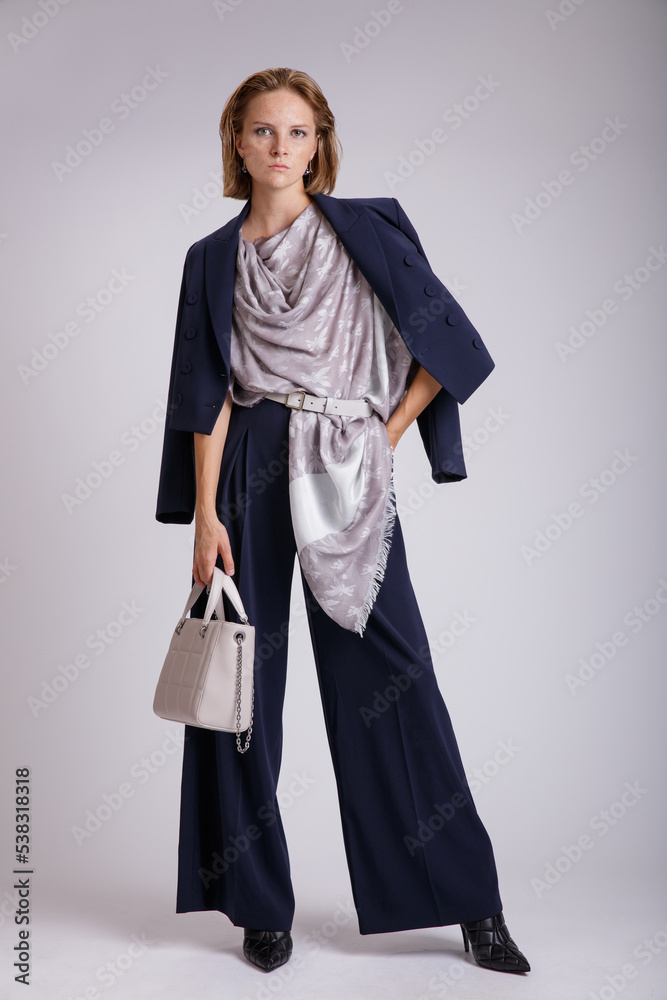 High fashion photo of a beautiful elegant young woman in a pretty blue suit, jacket, blazer, trousers, pants, handbag, scarf with a pattern posing over white, soft gray background. High fashion photo of a beautiful elegant young woman in a pretty blue suit, jacket, blazer, trousers, pants, handbag, scarf with a pattern posing over white, soft gray background.