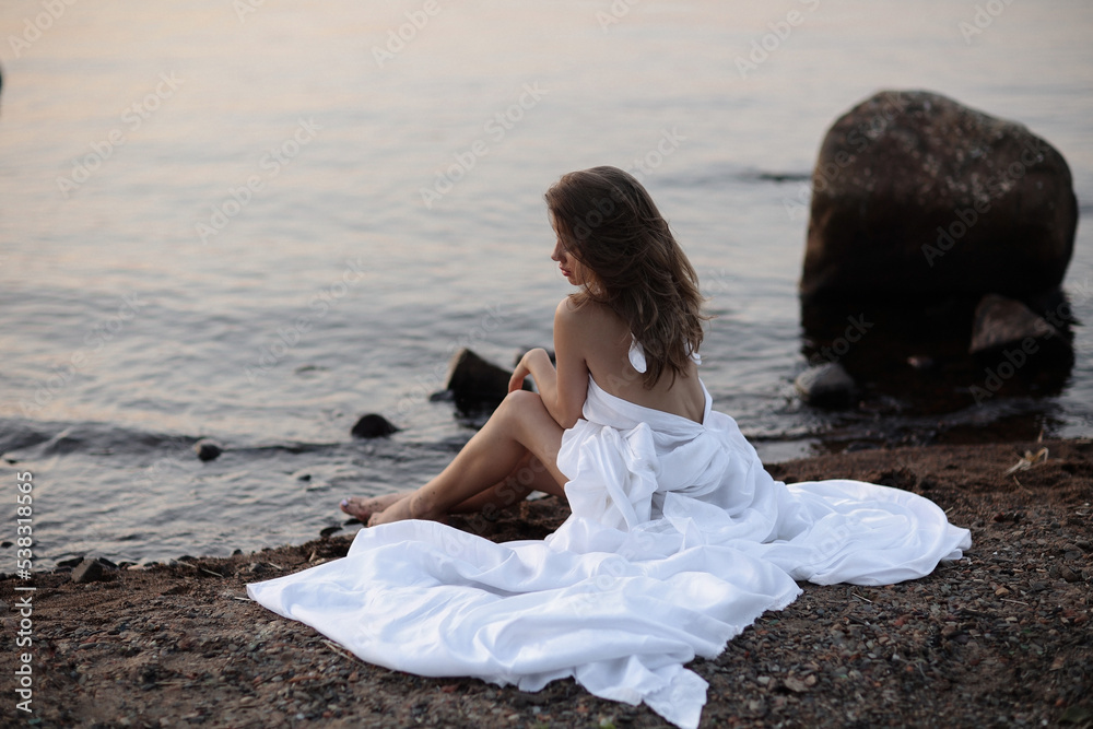 A beautiful brunette in a white dress sits on the shore. View from the back. A beautiful brunette in a white dress sits on the shore. View from the back.