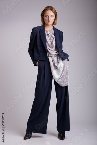 High fashion photo of a beautiful elegant young woman in a pretty blue suit, jacket, blazer, trousers, pants, scarf with a pattern posing over white, soft gray background. 