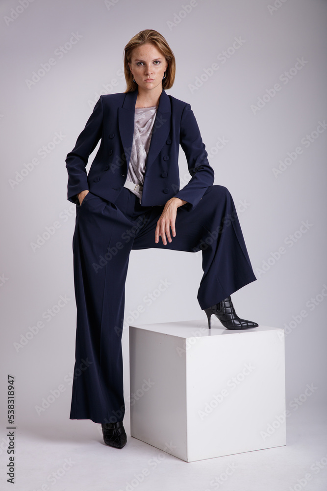 High fashion photo of a beautiful elegant young woman in a pretty blue suit, jacket, blazer, trousers, pants, scarf with a pattern posing over white, soft gray background. High fashion photo of a beautiful elegant young woman in a pretty blue suit, jacket, blazer, trousers, pants, scarf with a pattern posing over white, soft gray background.