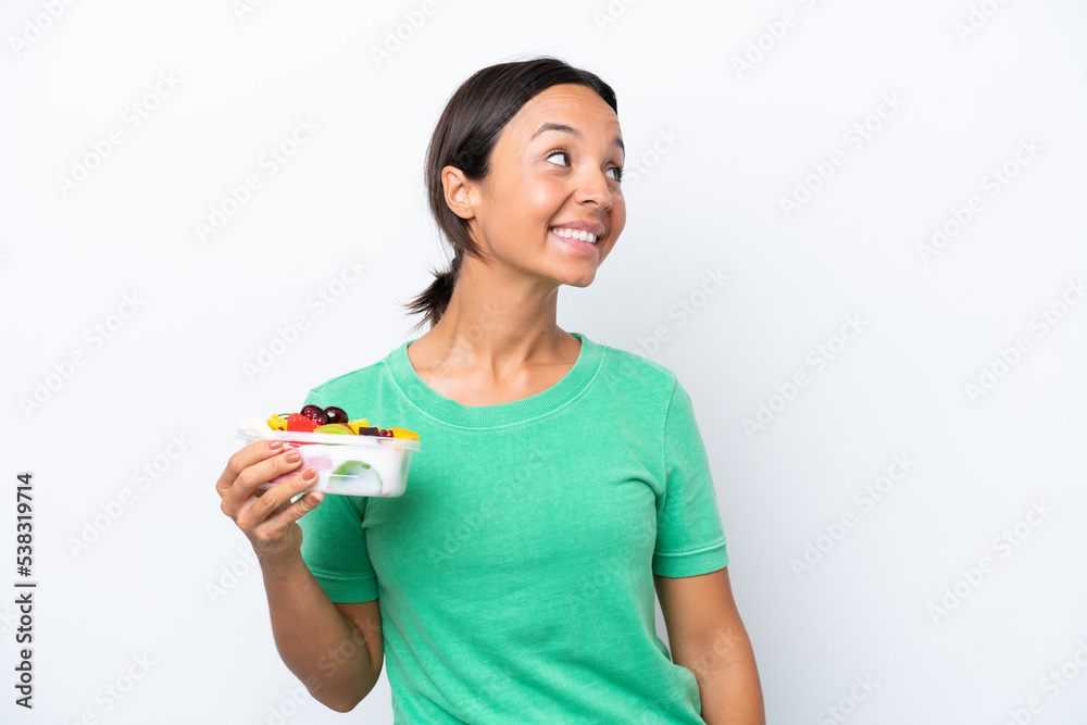 Young hispanic woman holding a bowl of fruit isolated on white background looking to the side and smiling Young hispanic woman holding a bowl of fruit isolated on white background looking to the side and smiling