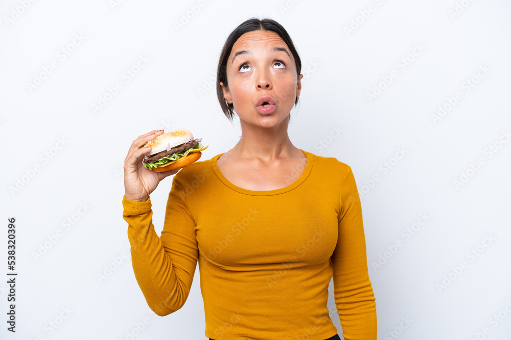 Young hispanic woman holding a burger isolated on white background looking up and with surprised expression Young hispanic woman holding a burger isolated on white background looking up and with surprised expression
