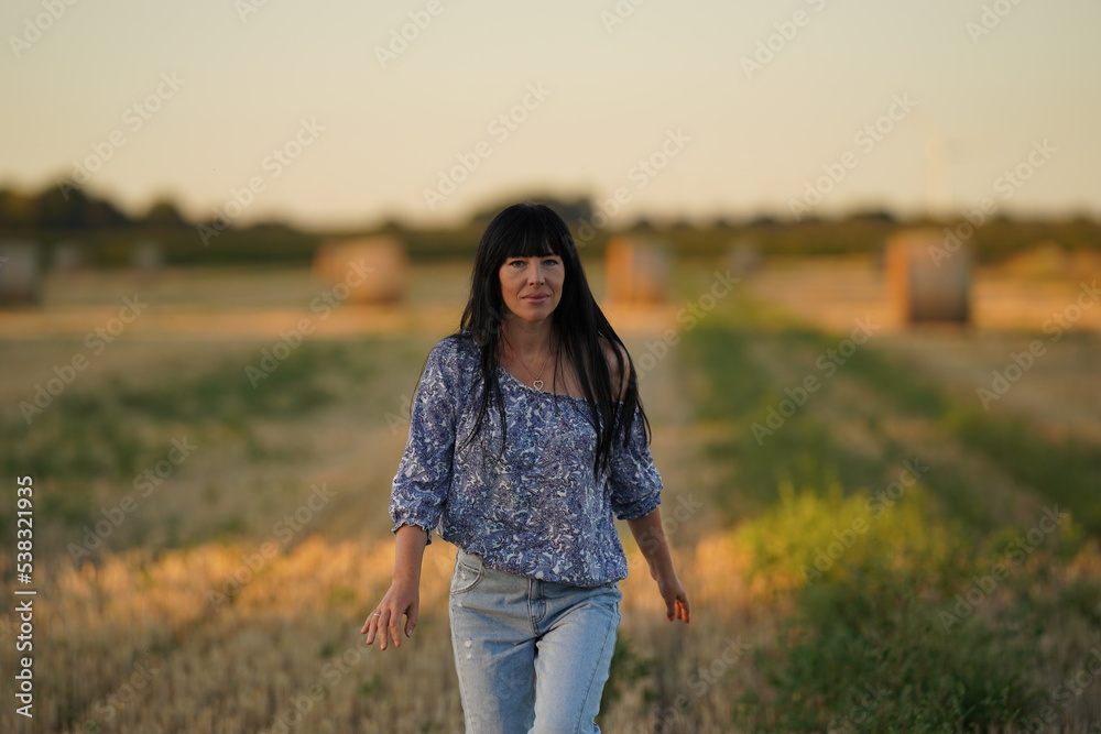 A girl with long black hair and a bare shoulder walks through a beveled field. She's wearing a light blouse and jeans. She is pleased and looks into the camera. Women's joy. A girl with long black hair and a bare shoulder walks through a beveled field. She's wearing a light blouse and jeans. She is pleased and looks into the camera. Women's joy.