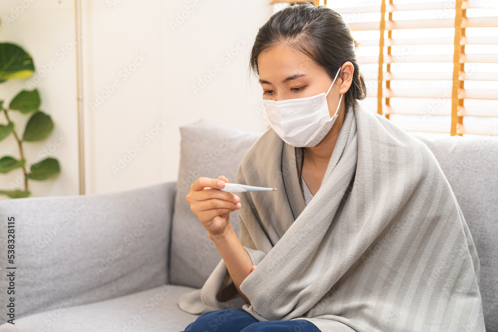 Sick, influenza asian young woman, girl wearing face mask in headache have a fever, flu and check thermometer measure body temperature, feel illness sitting on sofa bed at home. Health care, covid-19. Sick, influenza asian young woman, girl wearing face mask in headache have a fever, flu and check thermometer measure body temperature, feel illness sitting on sofa bed at home. Health care, covid-19.