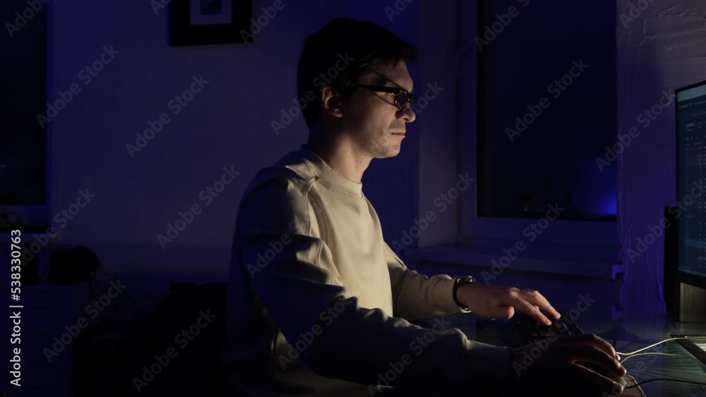 The male programmer works late. A guy with glasses is sitting in a dark office in front of a monitor and writing code. The male programmer works late. A guy with glasses is sitting in a dark office in front of a monitor and writing code.