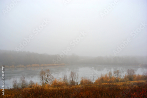 Beautiful evening morning, fog over lake, blue sky, trees in grass meadow, hills with yellow grass