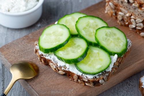 Slices of rye bread with cottage cheese and cucumbers on a wooden cutting board on grey background
