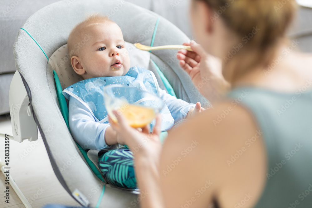 Mother spoon feeding her baby boy infant child in baby chair with fruit puree. Baby solid food introduction concept. Mother spoon feeding her baby boy infant child in baby chair with fruit puree. Baby solid food introduction concept.