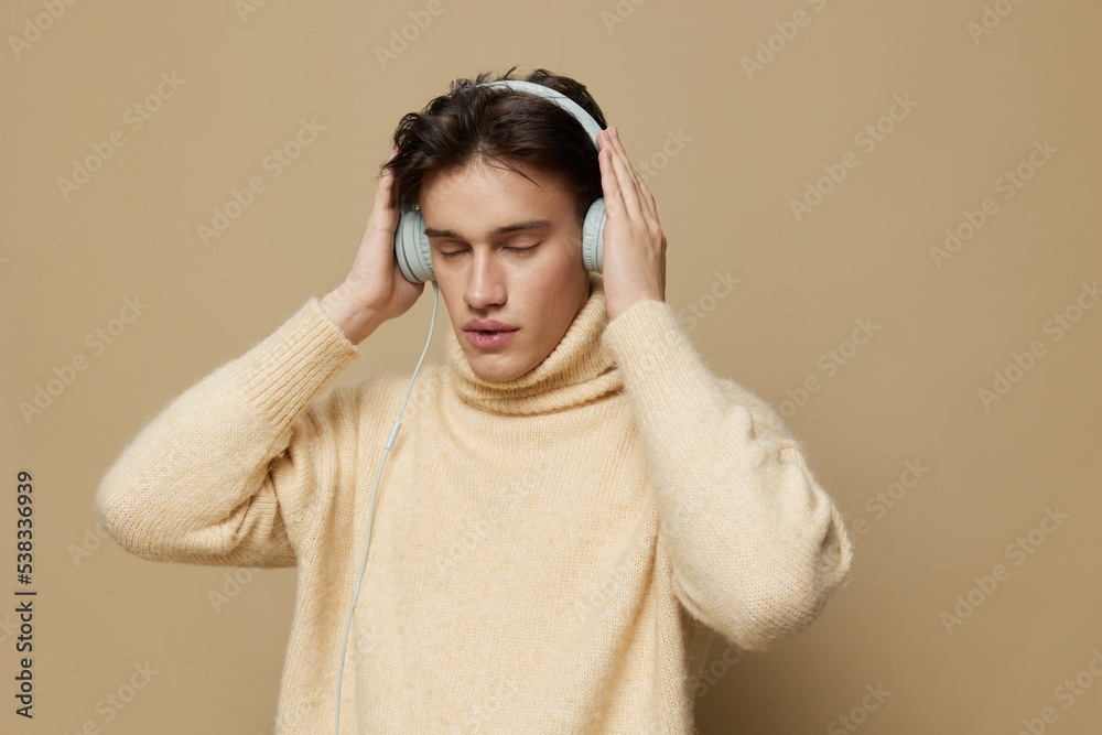 A attractive man with light white skin, with large headphones on his head with dark, short, short hair combed back, in a beige turtleneck with a high collar stands on a dark beige background.Close-up A attractive man with light white skin, with large headphones on his head with dark, short, short hair combed back, in a beige turtleneck with a high collar stands on a dark beige background.Close-up