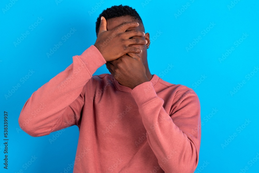 young handsome man wearing pink sweater over blue background Covering eyes and mouth with hands, surprised and shocked. Hiding emotions. young handsome man wearing pink sweater over blue background Covering eyes and mouth with hands, surprised and shocked. Hiding emotions.