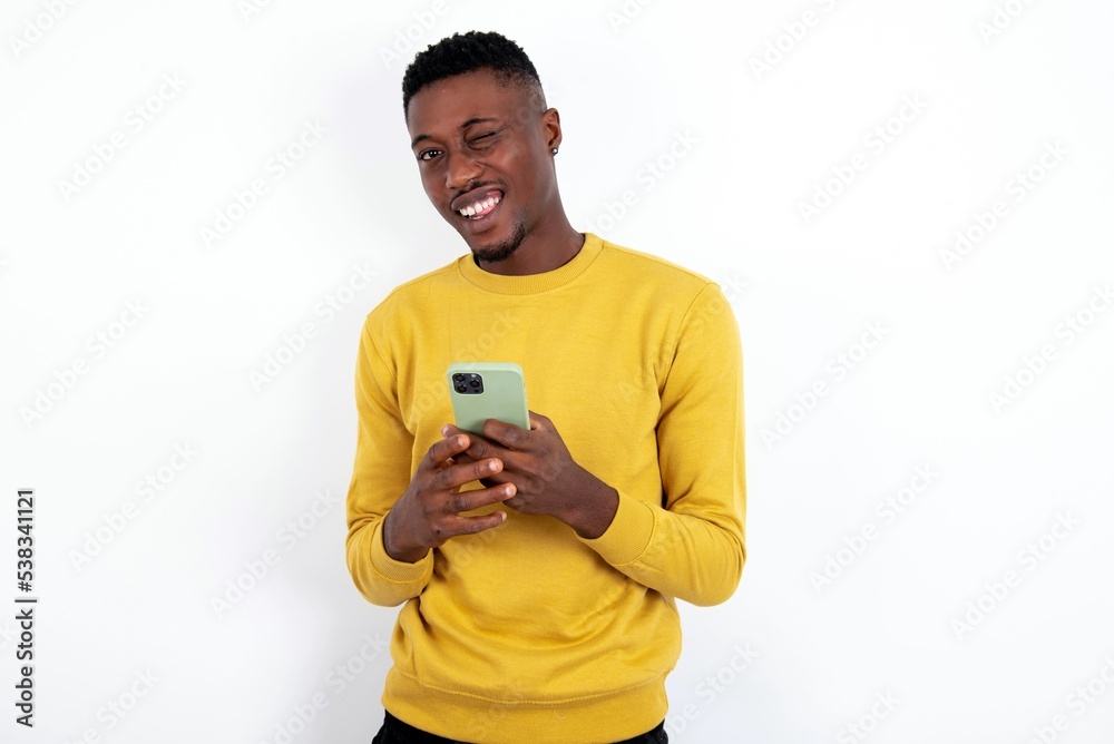 Pleased young handsome man wearing yellow sweater over white background using self phone and looking and winking at the camera. Flirt and coquettish concept. Pleased young handsome man wearing yellow sweater over white background using self phone and looking and winking at the camera. Flirt and coquettish concept.
