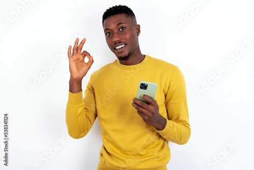 Happy young handsome man wearing yellow sweater over white background sending a message on his smartphone or taking a selfie and making ok sign with his hand.