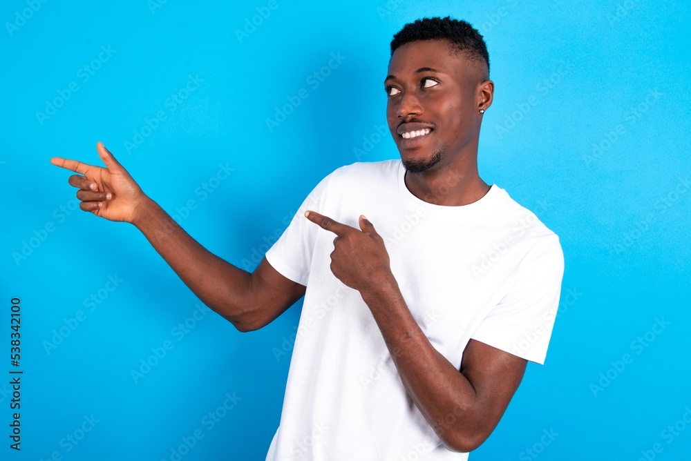 Optimistic young handsome man wearing white T-shirt over blue background points with both hands and looking at empty space. Optimistic young handsome man wearing white T-shirt over blue background points with both hands and looking at empty space.