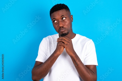 Curious young handsome man wearing white T-shirt over blue background keeps hands under chin bites lips and looks with interest aside.