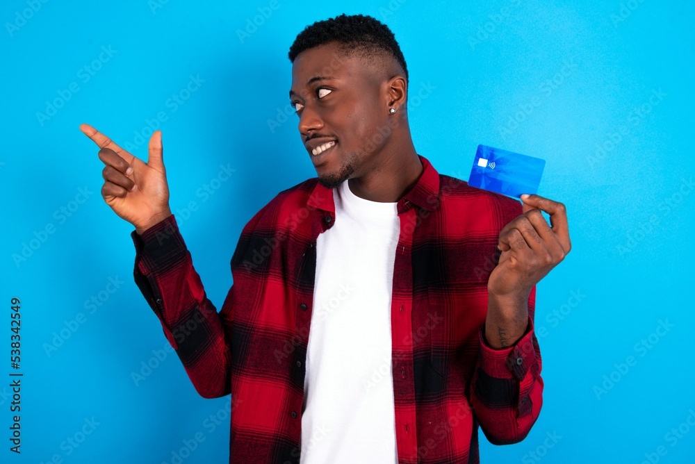 Curious smiling young handsome man wearing overshirt over blue background showing plastic bank showing finger copyspace Curious smiling young handsome man wearing overshirt over blue background showing plastic bank showing finger copyspace