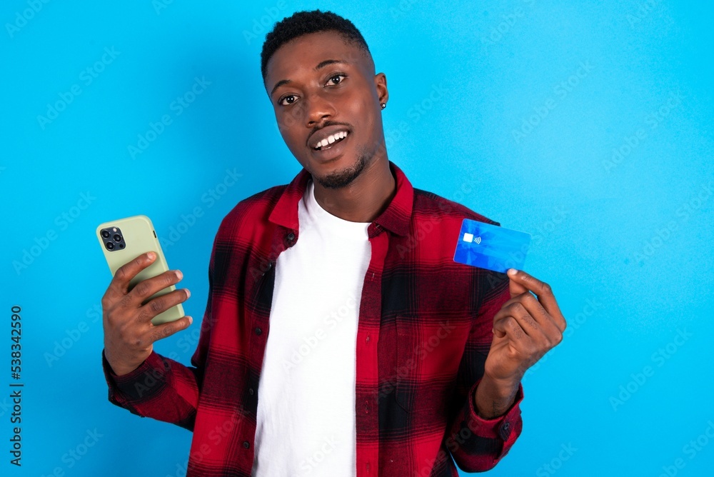 young handsome man wearing overshirt over blue background wears stereo headphones on ears holds modern mobile phone and credit card young handsome man wearing overshirt over blue background wears stereo headphones on ears holds modern mobile phone and credit card
