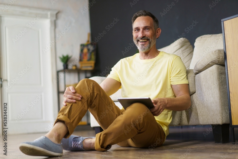 Handsome smiling man sitting on the floor with tablet pc in hand lean on sofa in living room have a rest. Businessman working. Business, education, lifestyle, interior design