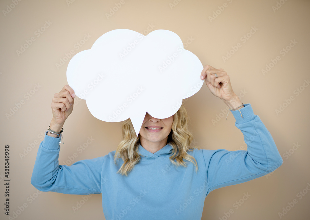 beautiful young woman with curly blond hair stands in front of brown wall and holds white speech bubble, thought bubble