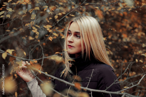 Blonde woman with long hair in a black jacket near a tree in the forest in autumn. Portrait of a girl with makeup in the autumn forest close-up.