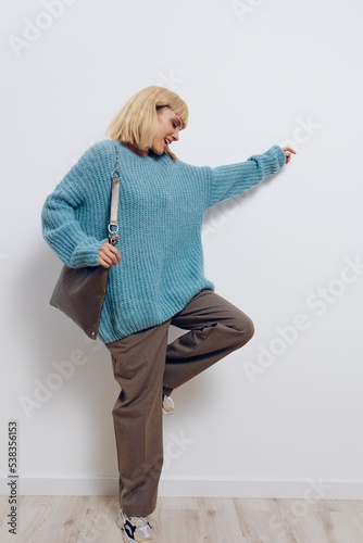 a full-length photo of a beautiful, happy, carefree stylish woman in a blue sweater and with a large bag, vigorously posing with her hand to the side