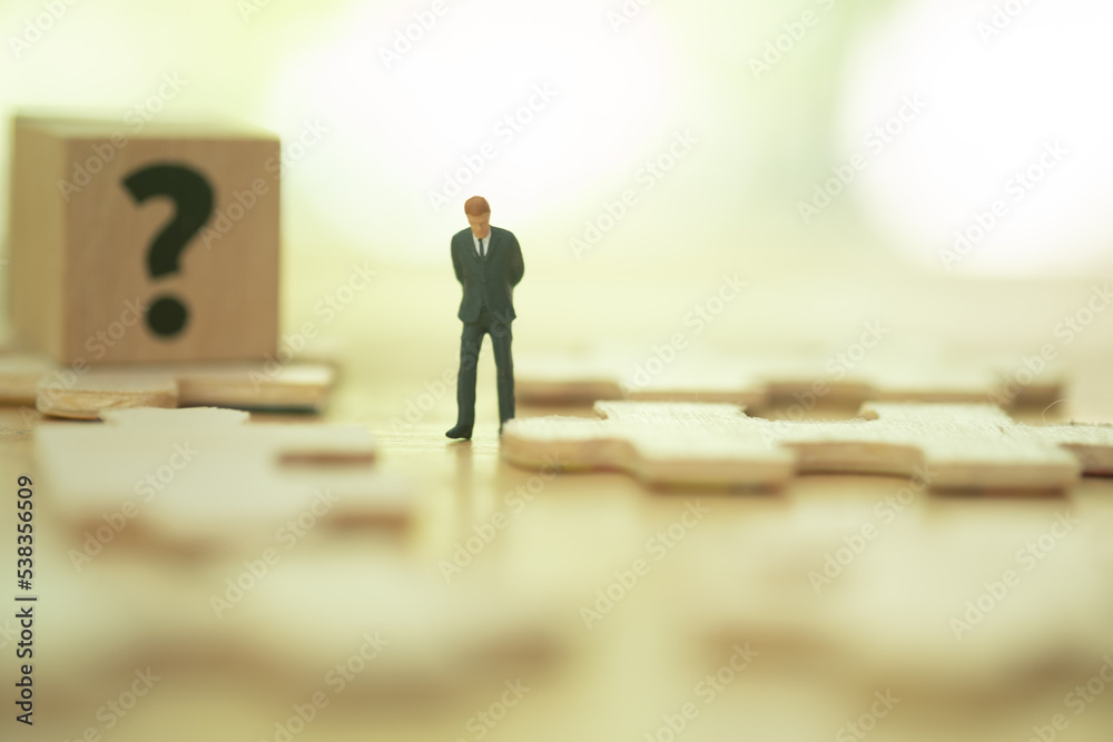 Fototapeta premium Miniature businessman in dark suit standing and thinking on wood block with jigsaw on floor and wood block with a question mark for thinking about business. Business and planning concept