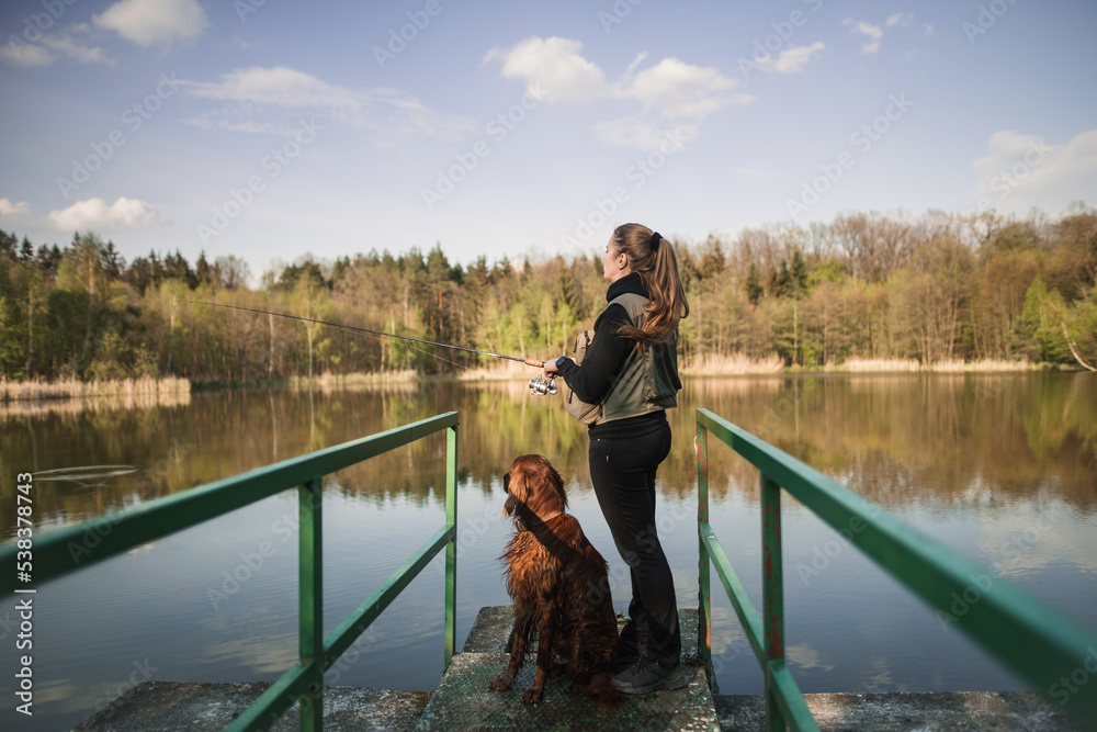 A young nice woman in a vest is fishing on a small pond with her hunting dog. Catching fish in nature at sunset. A relaxing hobby. 