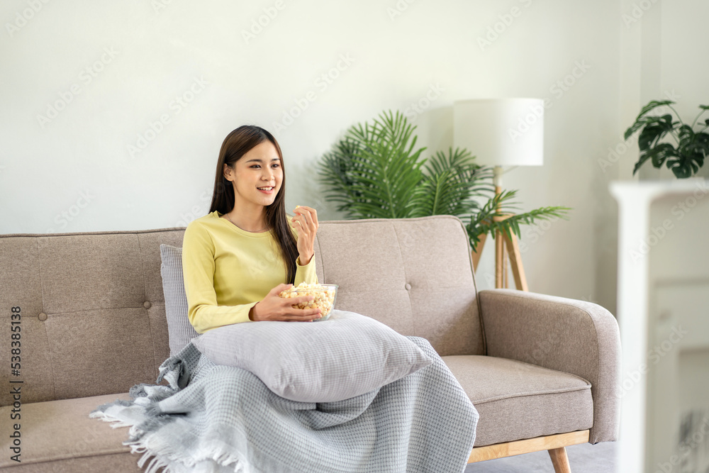 Young asian woman putting a bowl of popcorn on pillow and eating popcorn while sitting on the big comfortable Young asian woman putting a bowl of popcorn on pillow and eating popcorn while sitting on the big comfortable